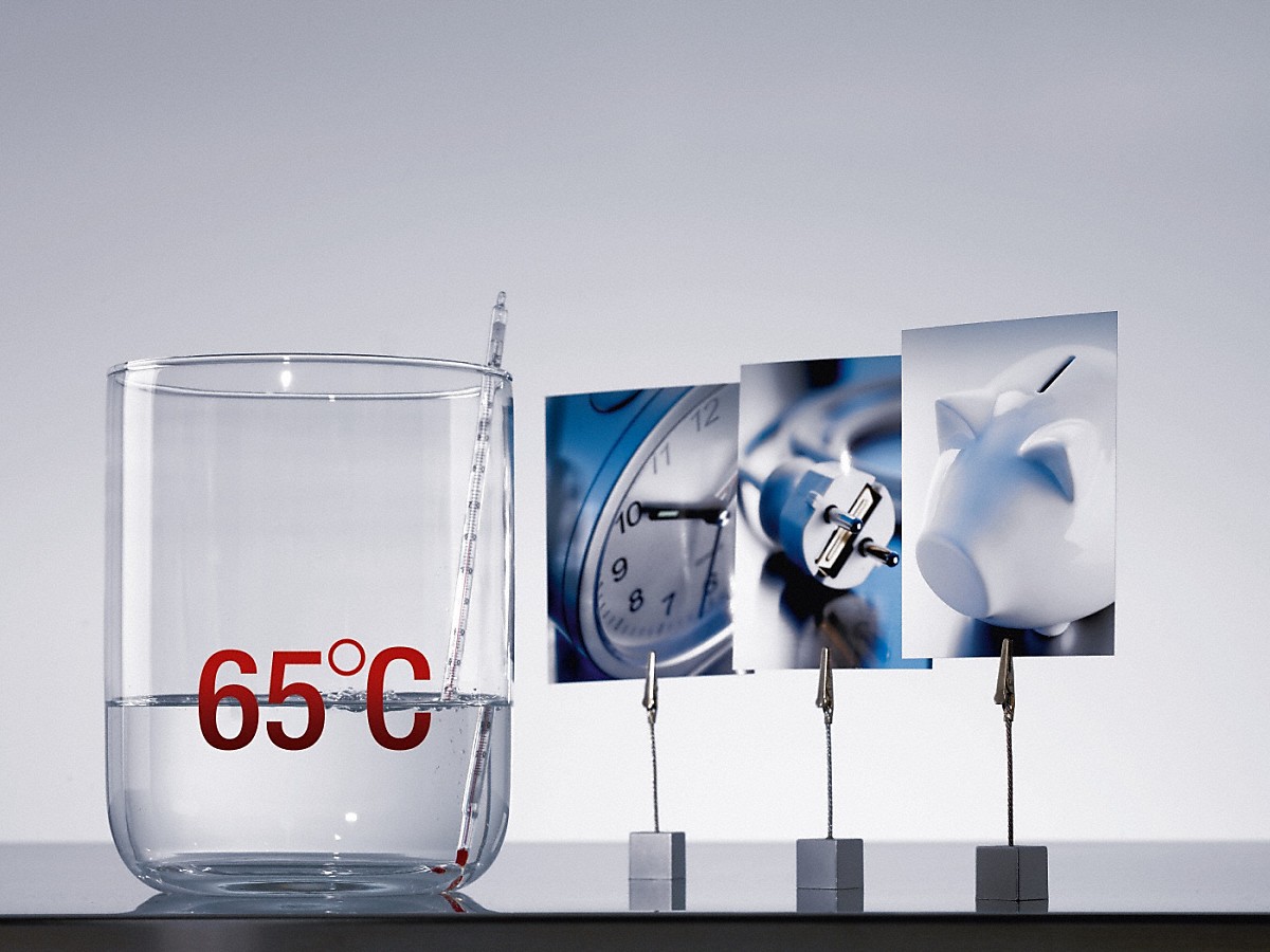 Miele Warmwasseranschluss Geschirrspüler etwas wasser in einem glas mit roter 65°C aufdruck neben drei Fotoklipphaltern mit einer Uhr, einem Steckdosenkabel und einem Sparschwein vor einer hellgrauen Wand