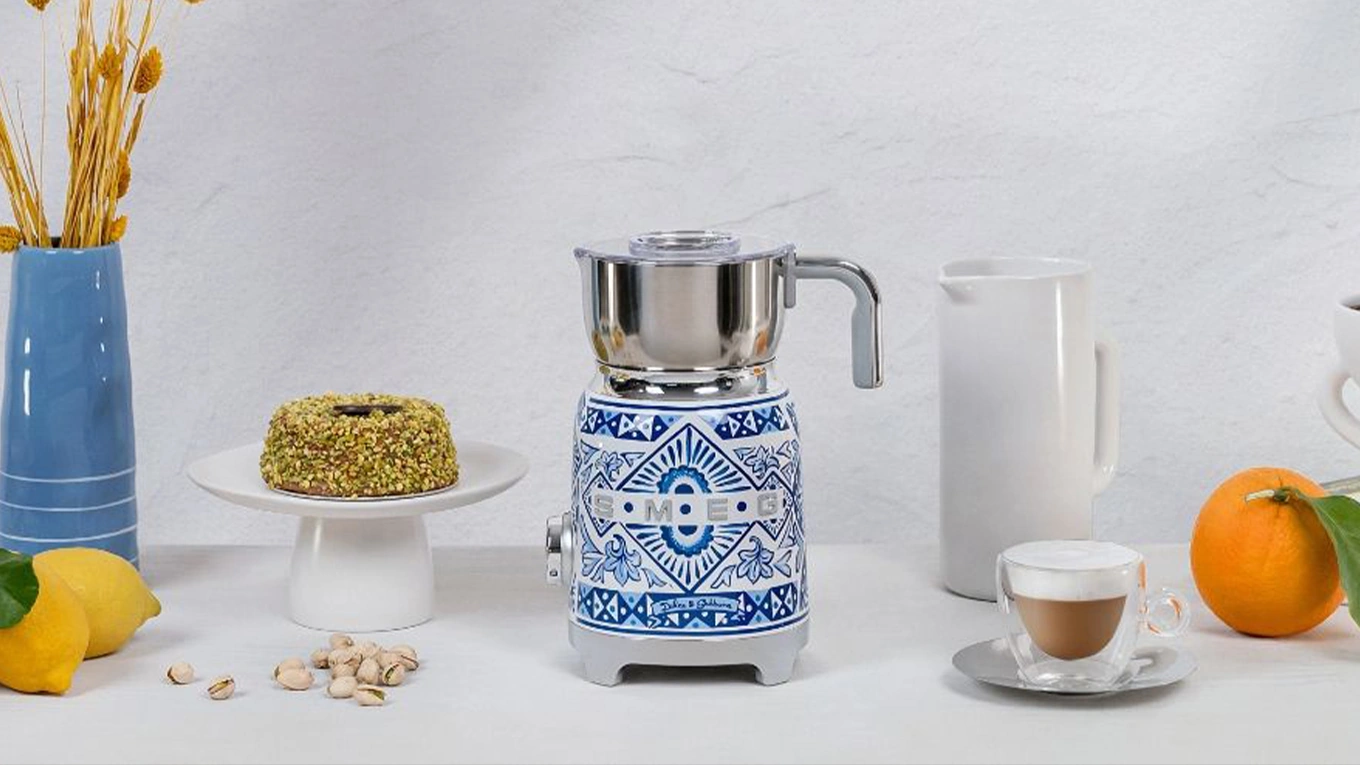 smeg dolce & gabbana blue mediterraneo milchaufschäumer in blau-weiß mediterran stil neben einem milchkaffee und kuchen