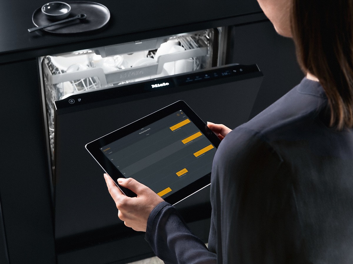 Miele Autostart Geschirrspüler tablett mit gelben balken auf dem display in den haenden einer frau vor einem halb geoeffneten Miele Geschirrspueler vollintegriert in einer dunklen kueche