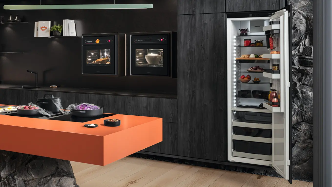 bora kühlschrank mit boxen in einer dunklen küche neben backofen und hinter einer kücheninsel mit kochfeldabzug