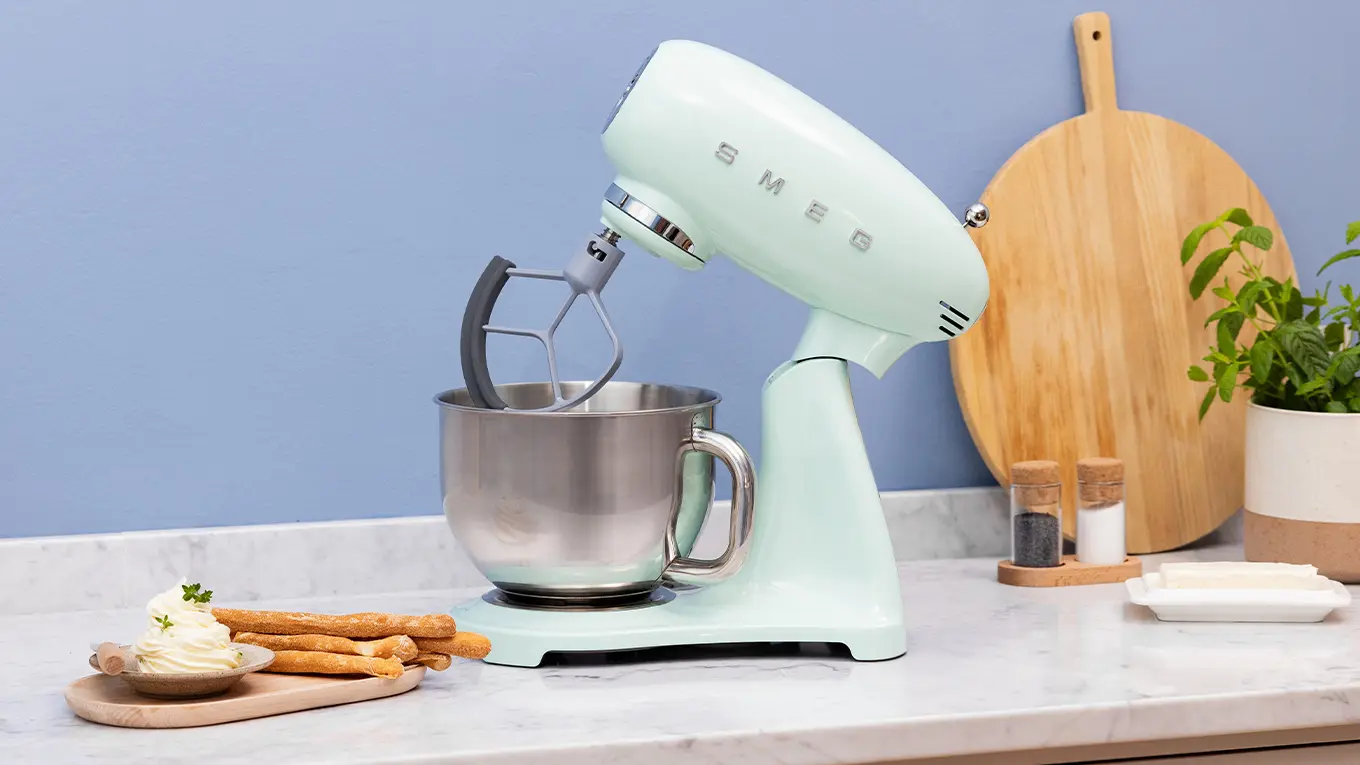 smeg küchenmaschine smf05 in pastellgrün auf einer marmor arbeitsplatte neben holzbrett und servierteller