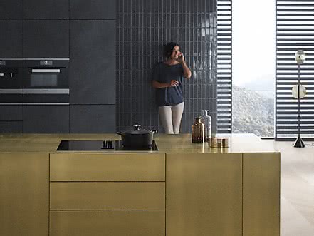 Miele KMDA Kochfeld in einer goldenen Beispielkueche mit telefonierender Frau im Hintergrund