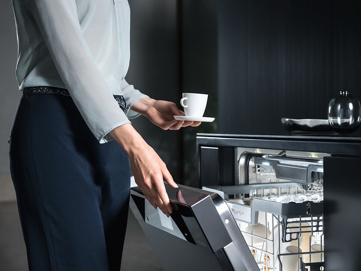 Miele Comfort Close Geschirrspüler frau mit einer kleinen weissen kaffeetasse mit untertasse in der hand beim oeffnen von einem miele geschirrspueler
