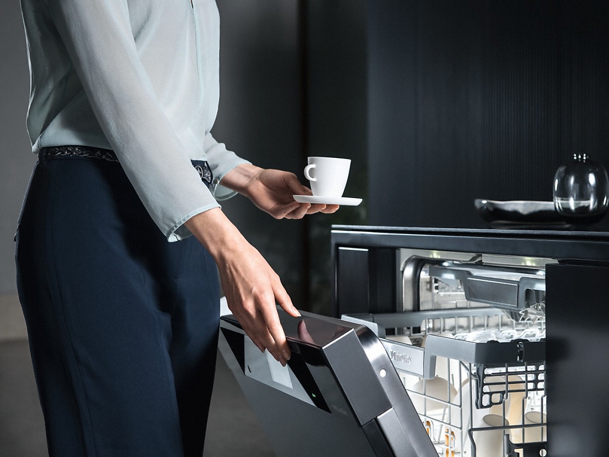 frau mit einer kleinen weissen kaffeetasse mit untertasse in der hand beim oeffnen von einem miele geschirrspueler