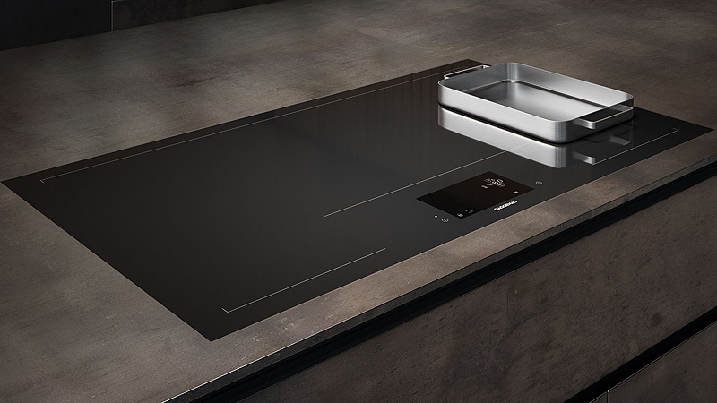 gaggenau serie 400 induktionskochfeld mit flächeninduktion auf dem ein edelstahlbraeter rechts steht von der seite in einer metallic kuechenplatte