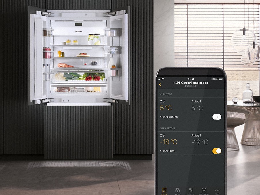 smartphone mit temperaturanzeige für kühl-gefrierkombination aus der miele app auf dem display vor einem geöffneten side by side kühlschrank in einer wohnküche
